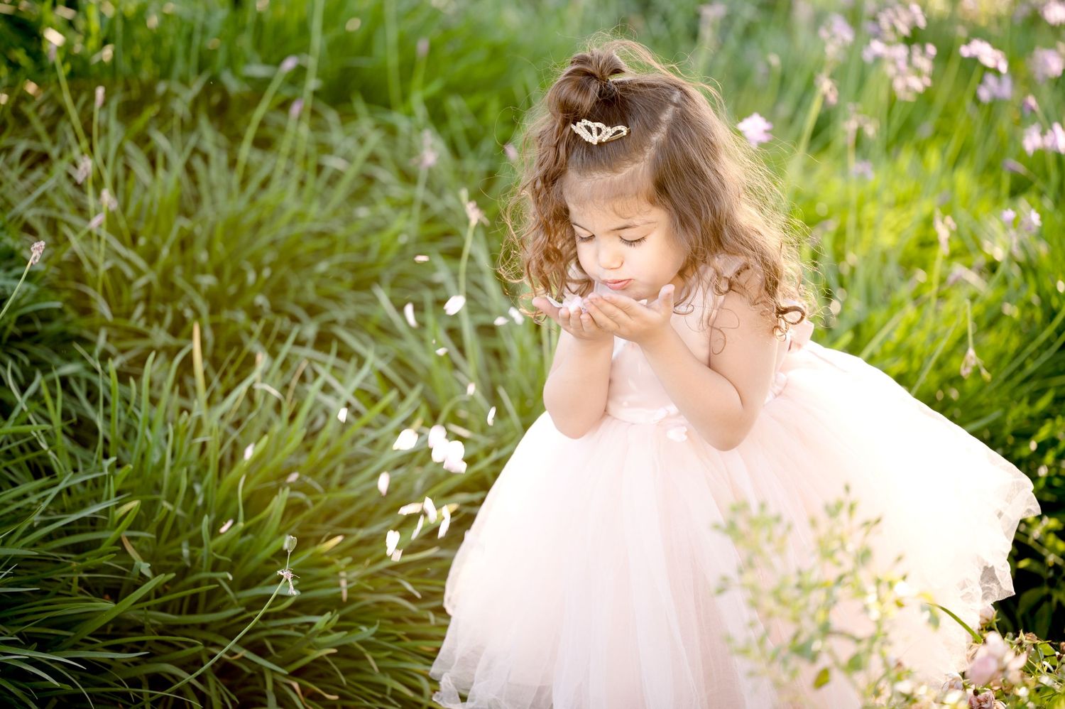 A small figure in a pink tulle dress giggles while playing in a sun-lit garden with tall grass and flowers.