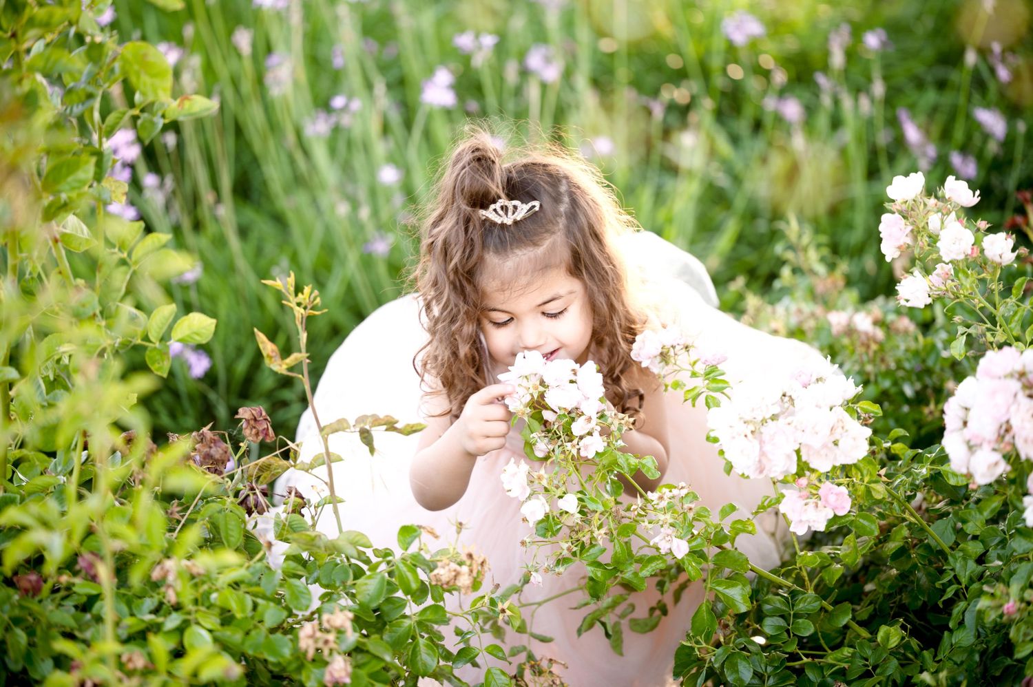 A whimsical scene shows a figure in a pink dress bending down to smell white flowers in a garden.