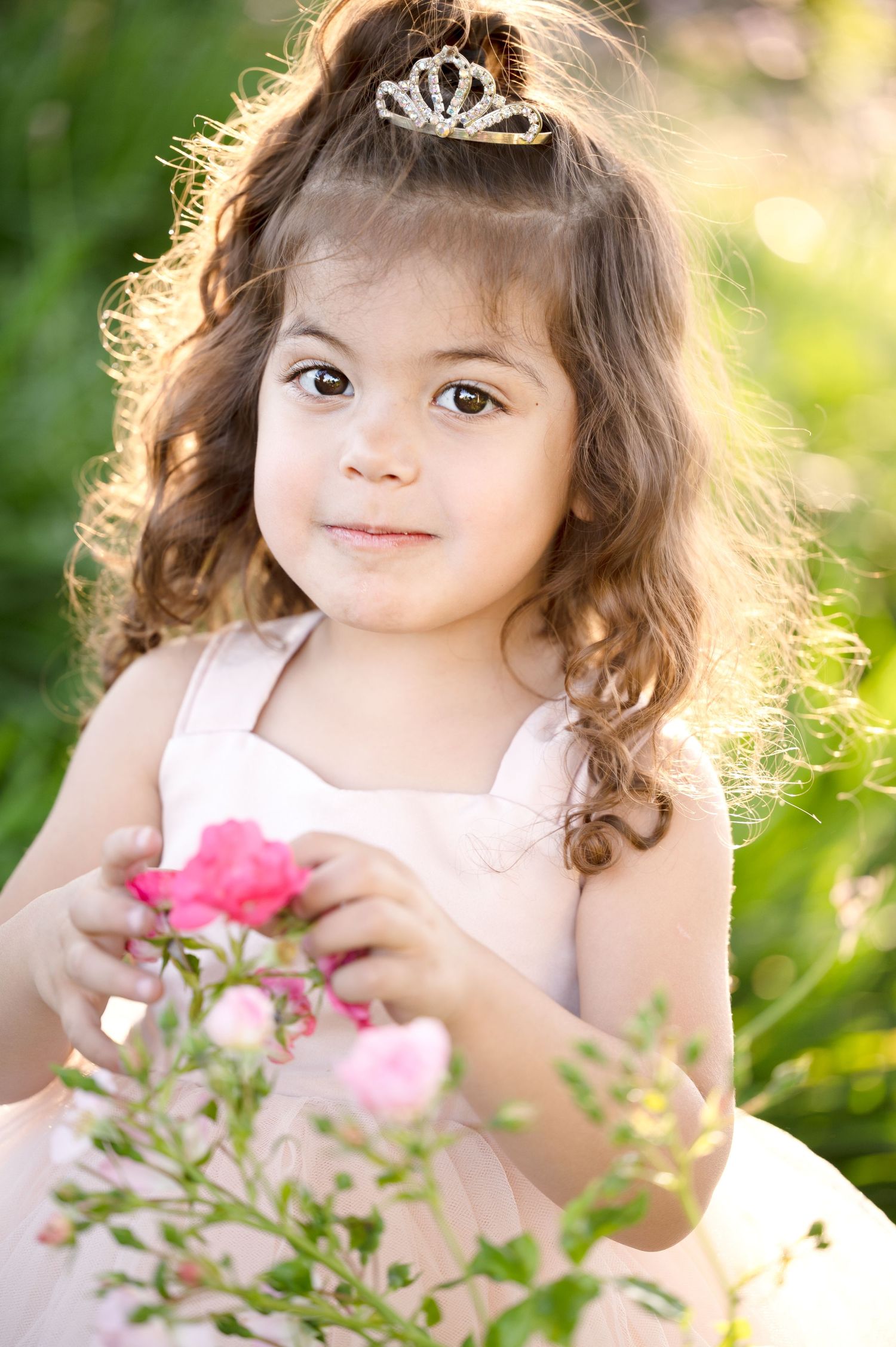 A portrait in a garden setting features a tiara and pink dress against natural sunlit greenery.