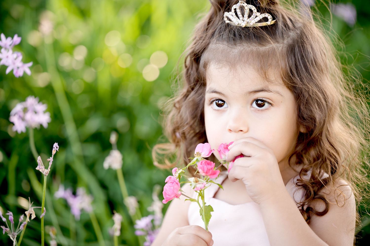 A dreamy garden portrait shows someone smelling pink flowers while wearing a tiara and dress.