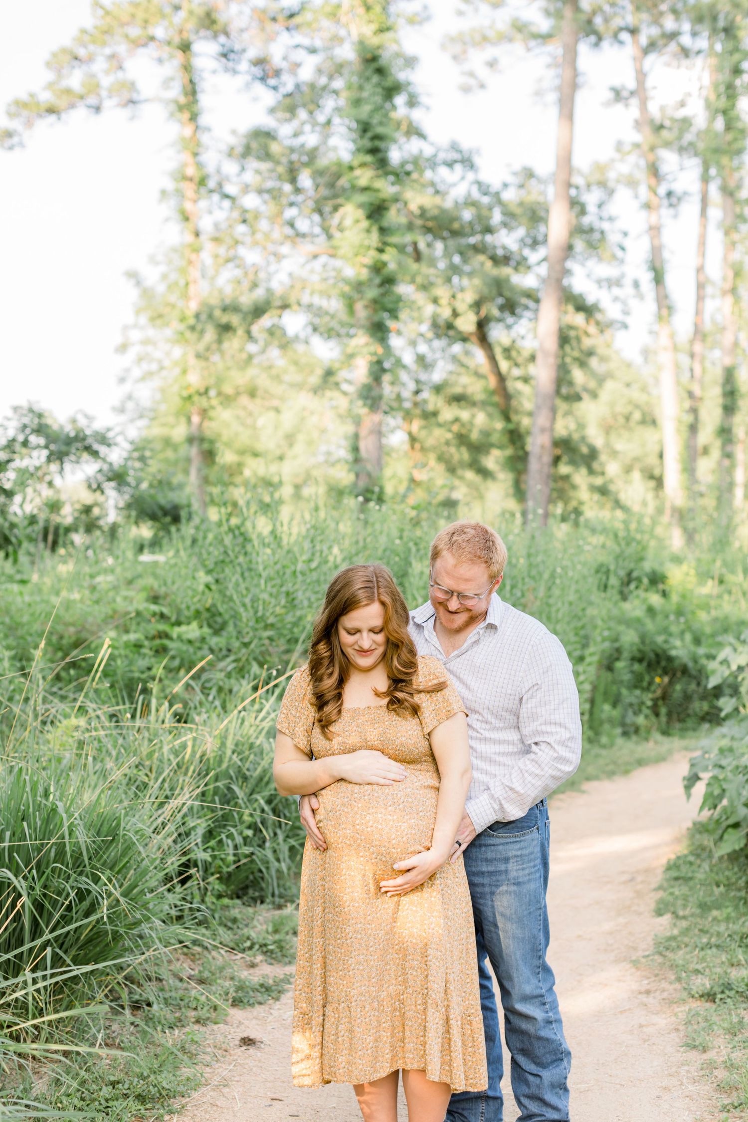 Couple stands together on nature path with woman in yellow floral dress cradling baby bump.