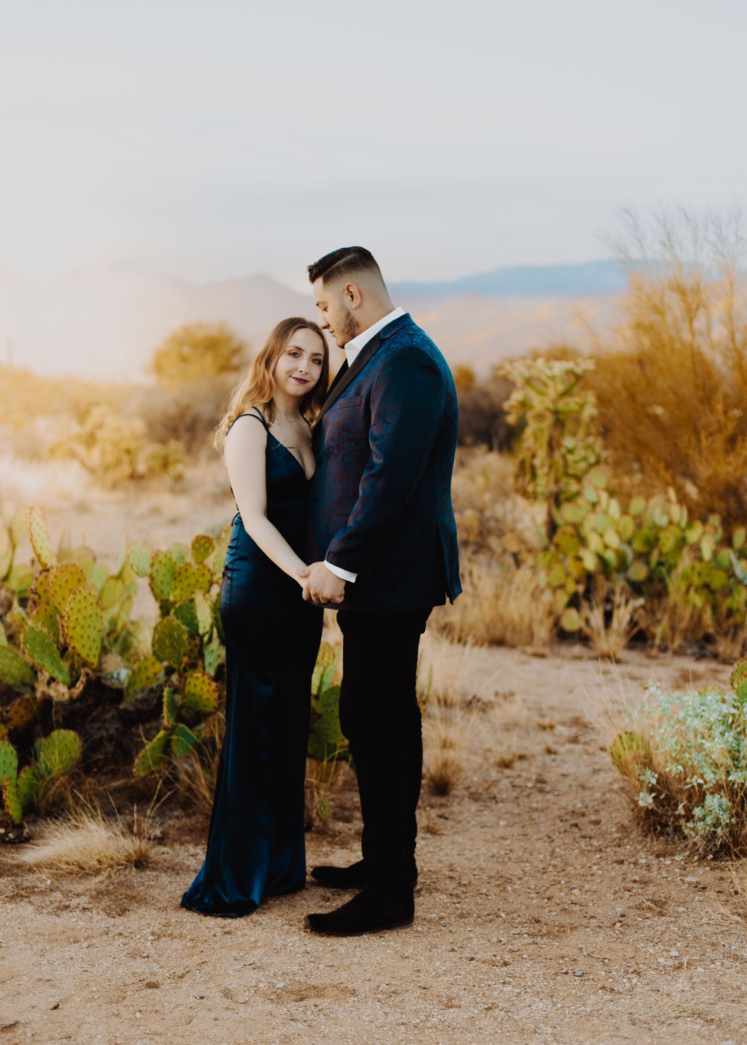 Couple in formal attire embracing on desert path at sunset.