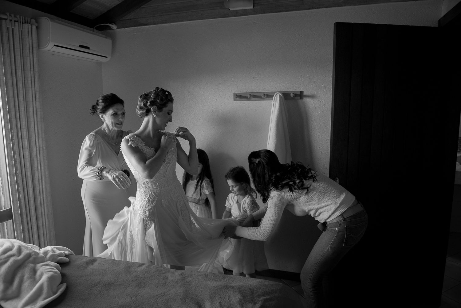 Black and white photo capturing wedding dress preparation in an intimate indoor setting.