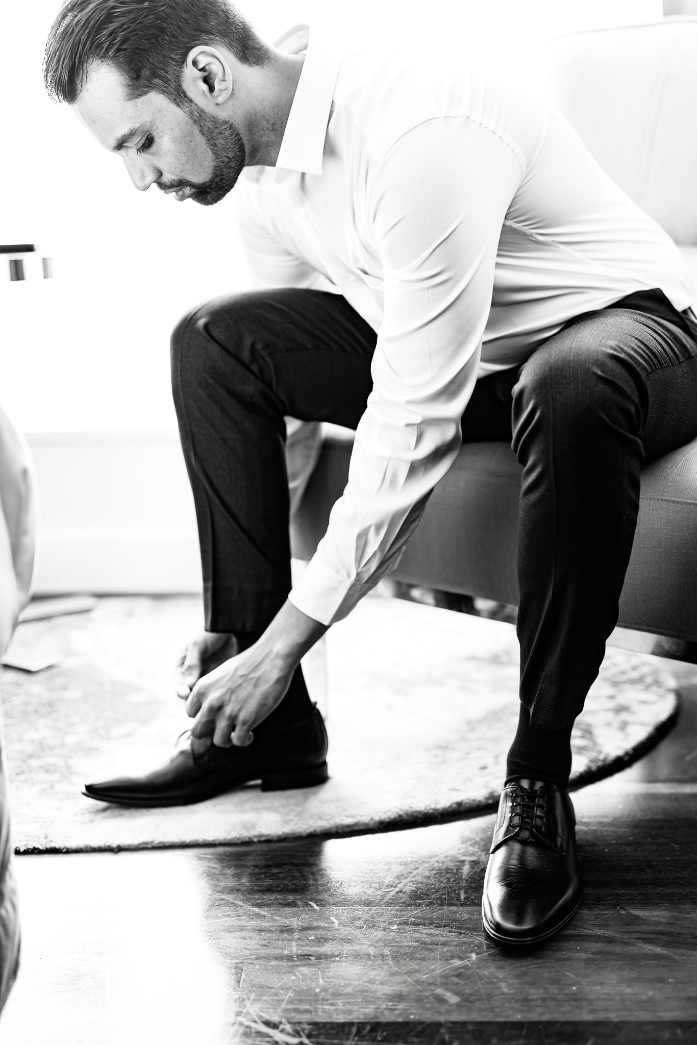 A black and white photo showing formal dress shoes being put on while seated on a white couch.