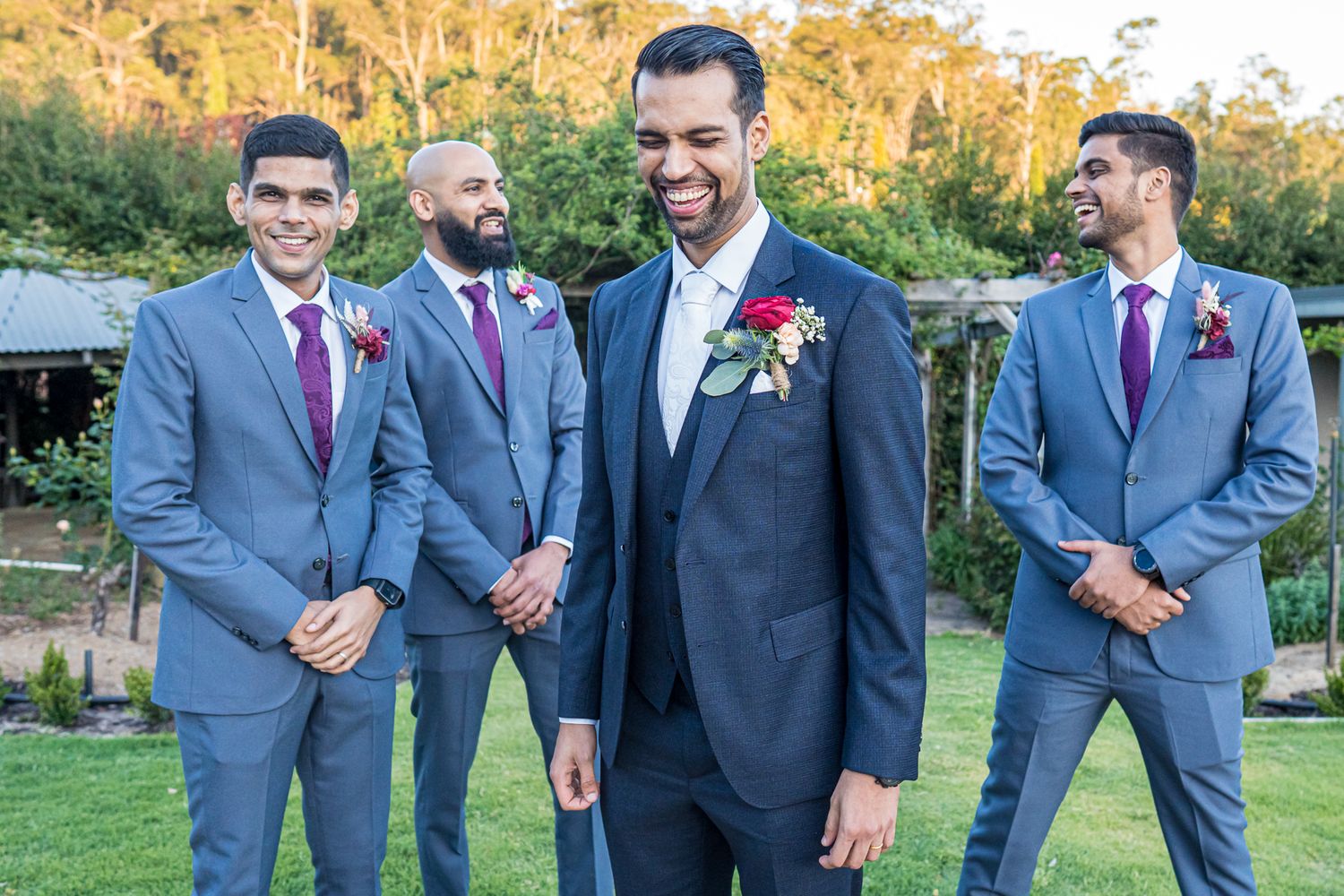 Groomsmen in gray and navy suits with burgundy ties pose outdoors in natural lighting.