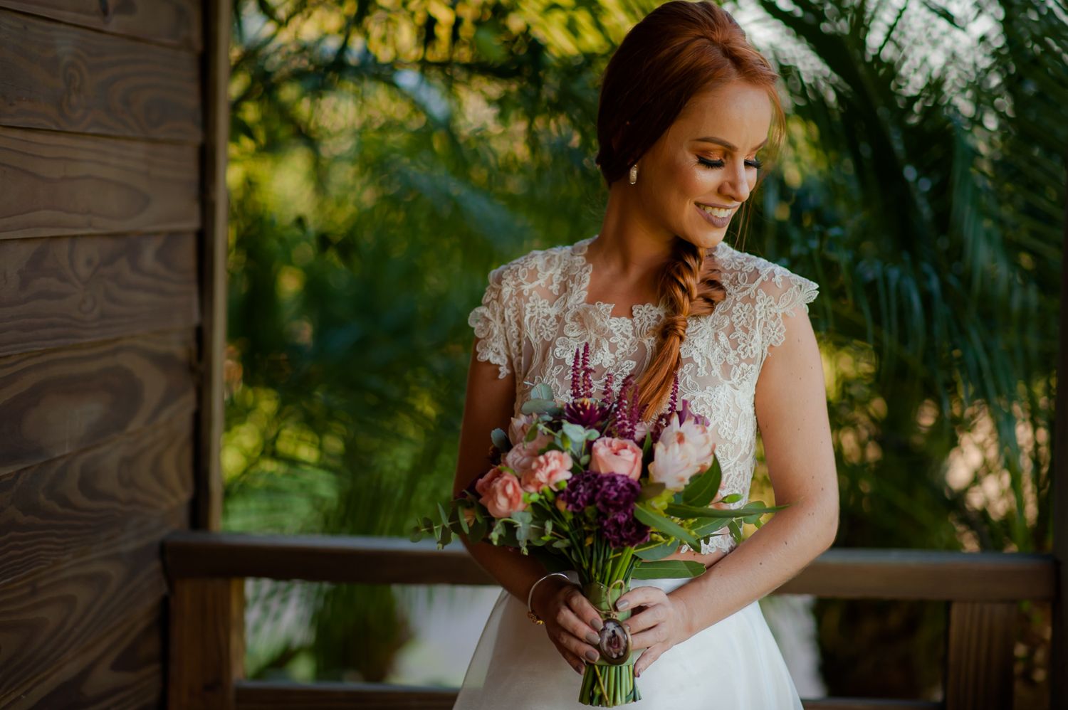 Bride in lace dress holding colorful bouquet while standing in rustic setting.