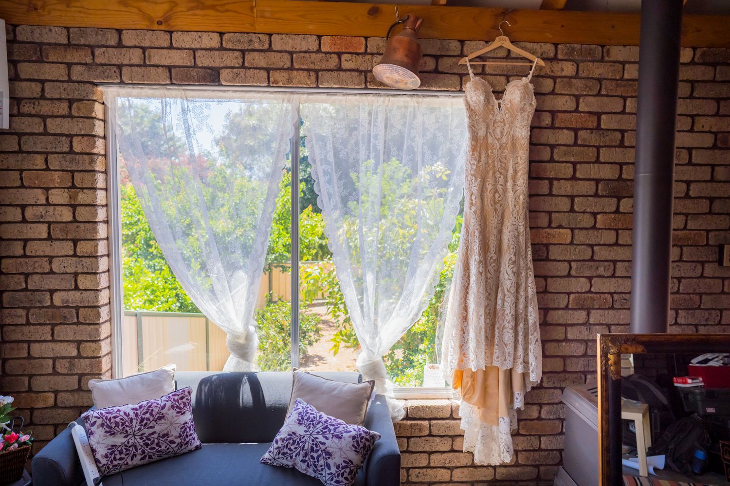 A lace wedding dress hangs by a window with white curtains and brick walls.
