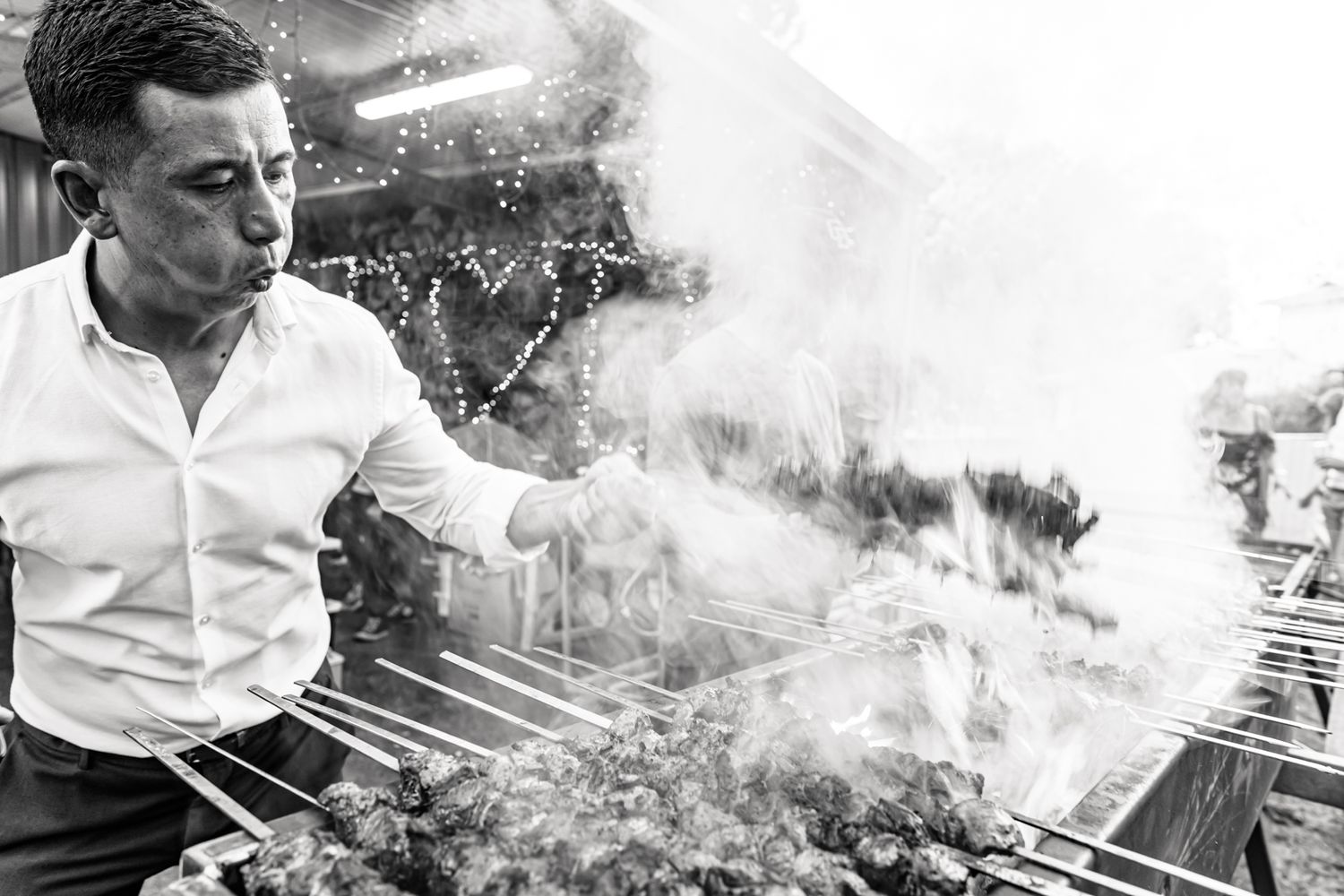 Chef in white shirt grilling food with dramatic smoke rising from outdoor barbecue in black and white photo.