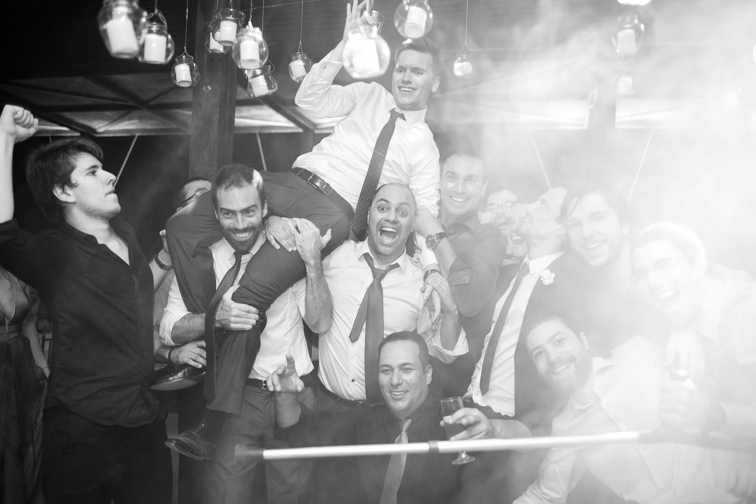 Black and white photo of celebratory crowd at wedding reception with bright lights above.