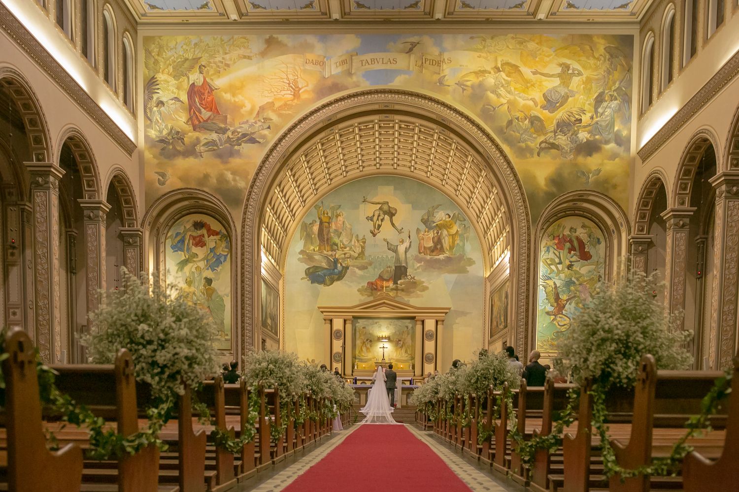 A grand church interior with ornate ceiling murals, arched columns and floral arrangements lining the center aisle.