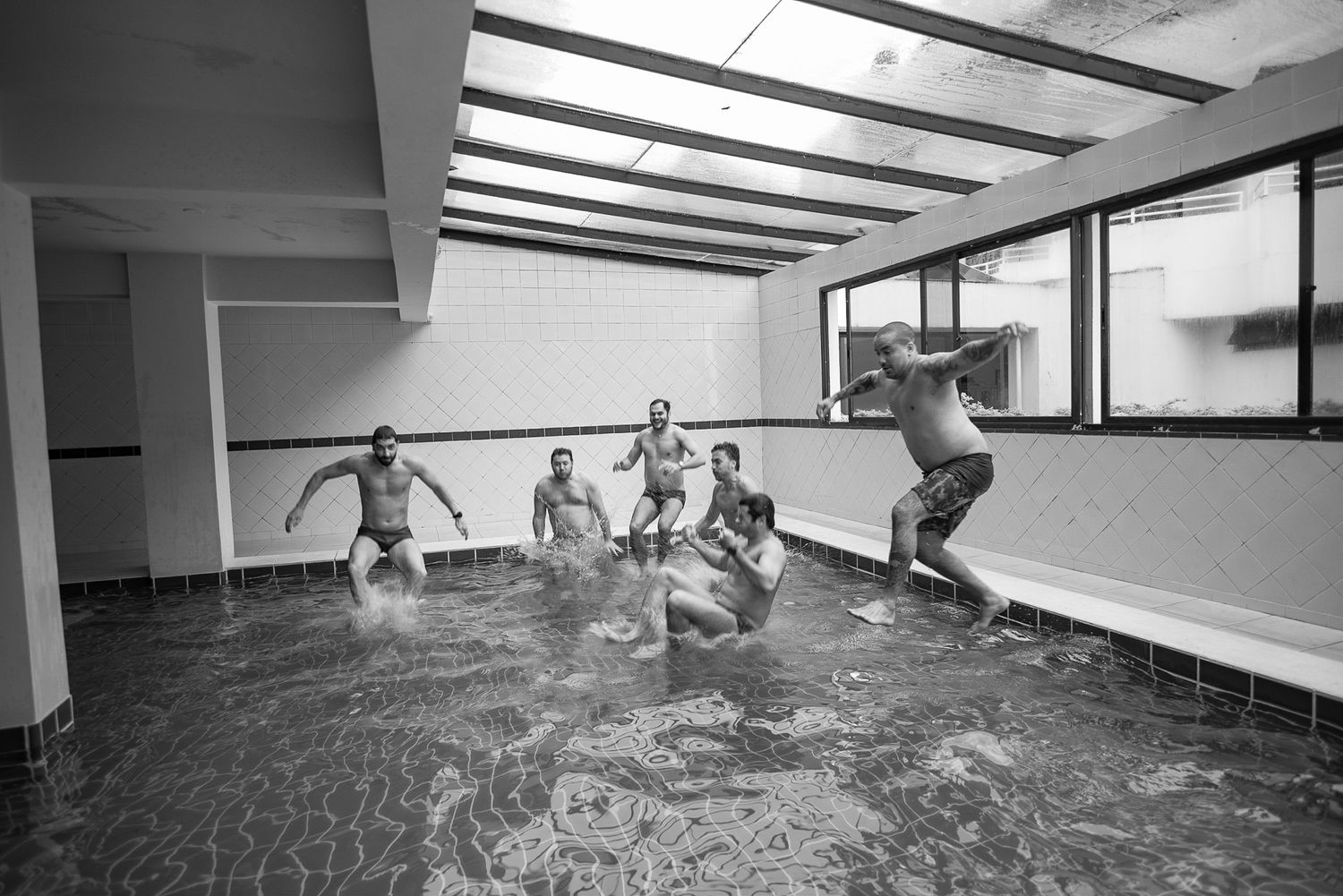 Joyful people jumping into an indoor swimming pool creating splashes in black and white photography.