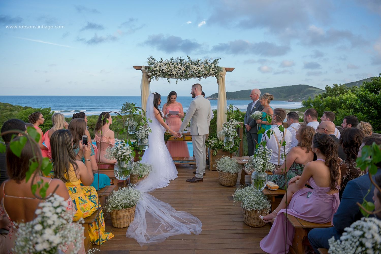 Outdoor wedding ceremony on seaside deck with floral arch and gathering of guests.