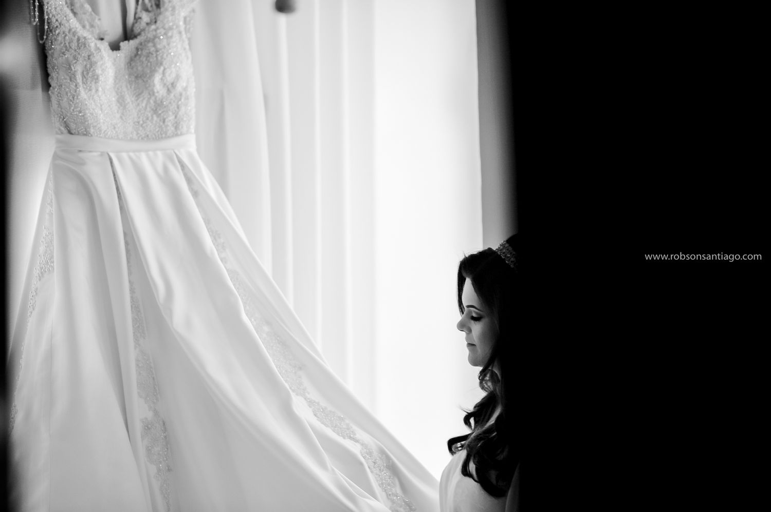 A wedding dress hangs elegantly by a window in black and white photography.