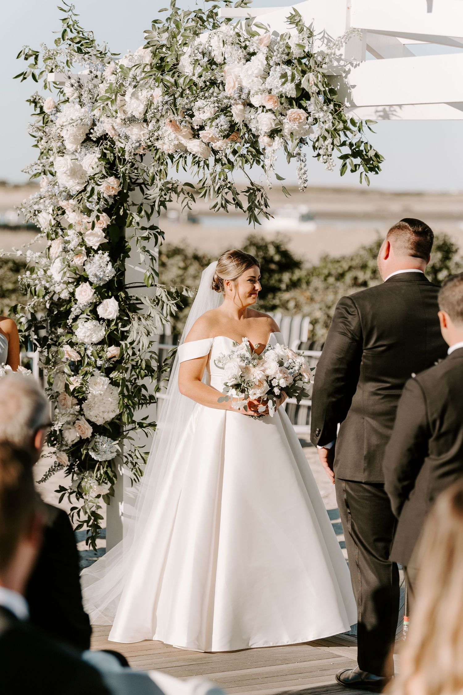 A wedding ceremony under a white pergola adorned with lush floral arrangements.