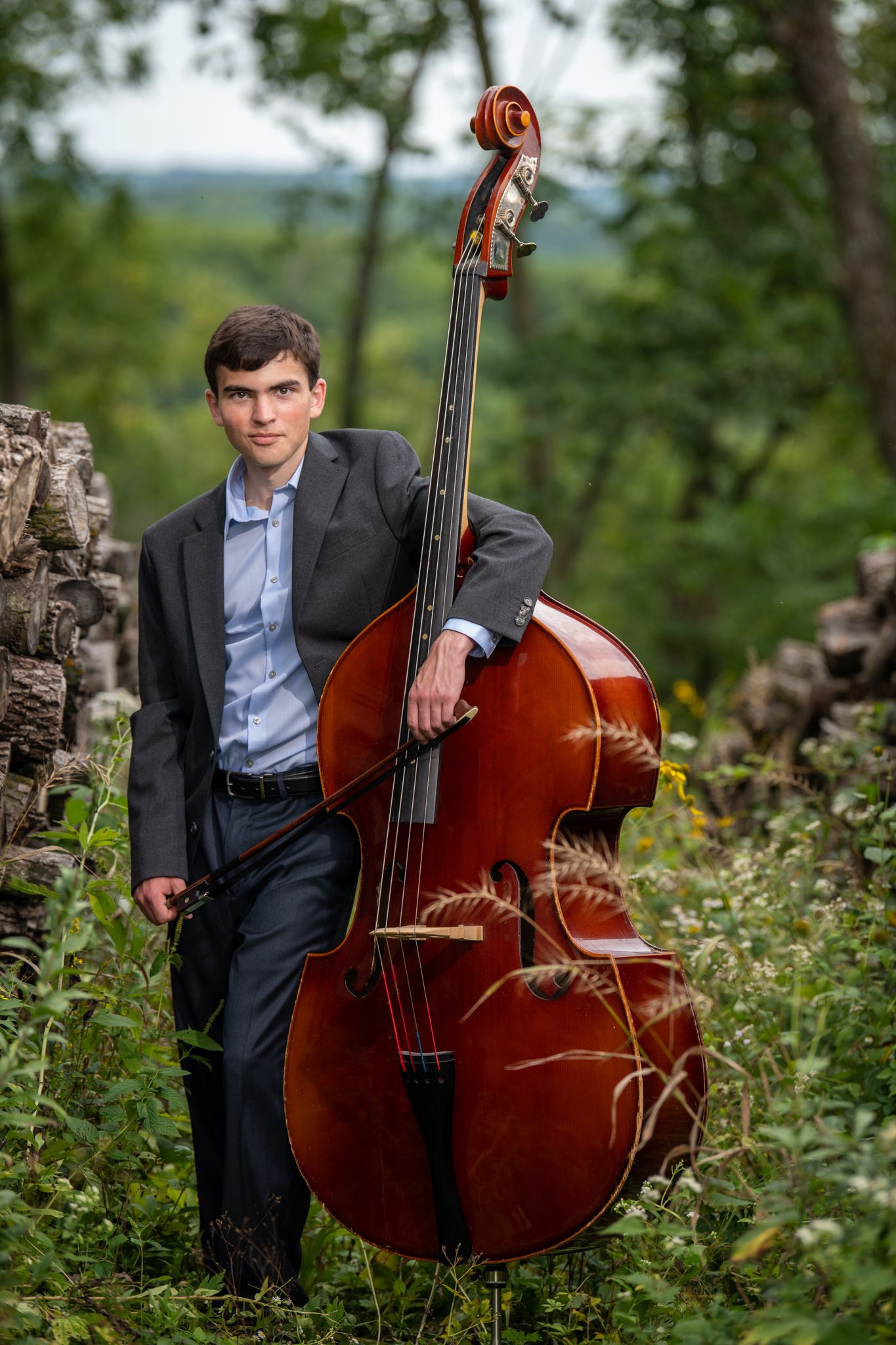 Musician poses with upright bass instrument in outdoor nature setting with trees in background.