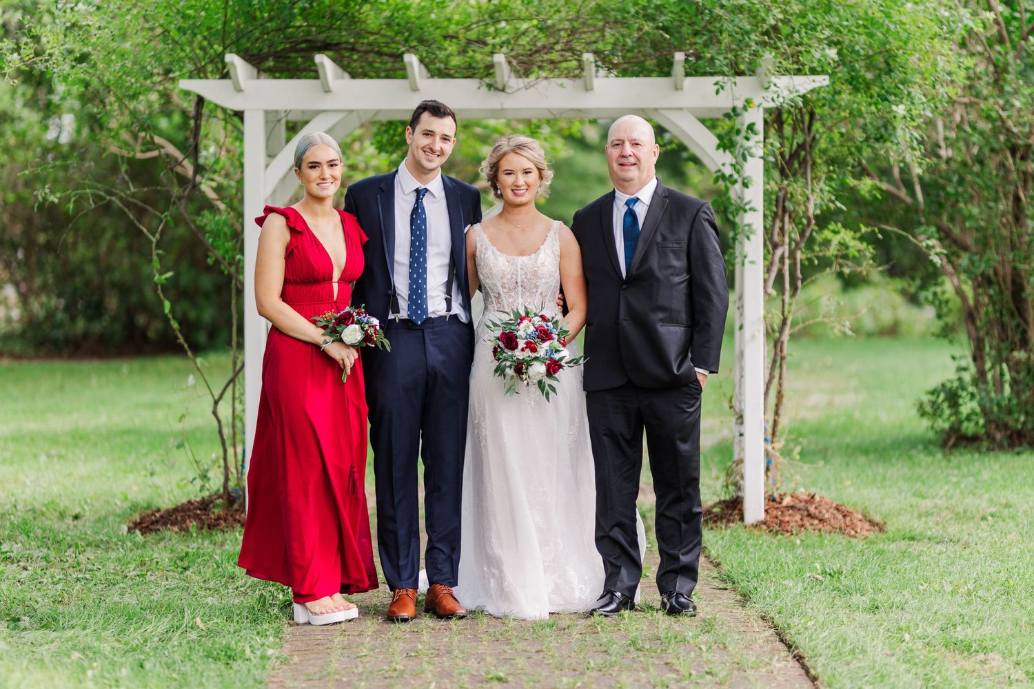 Small wedding group posing under garden pergola with bride in white and guest in red dress.