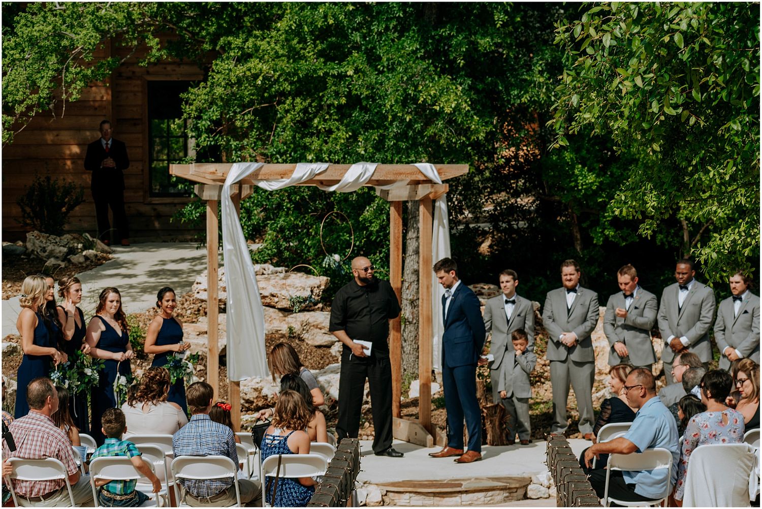A rustic outdoor wedding ceremony setup with a wooden arch and white fabric draping surrounded by greenery.