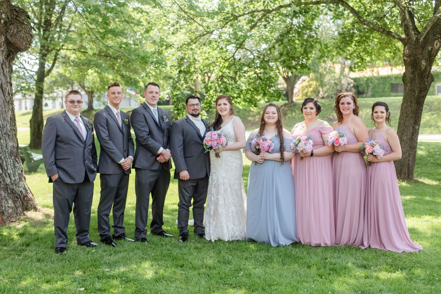Wedding party in pastel dresses and dark suits poses outdoors under green trees.