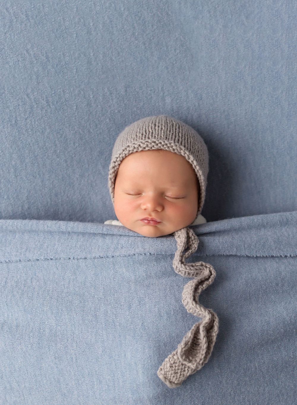 Sleeping newborn wearing knit bonnet with long tail against blue background.