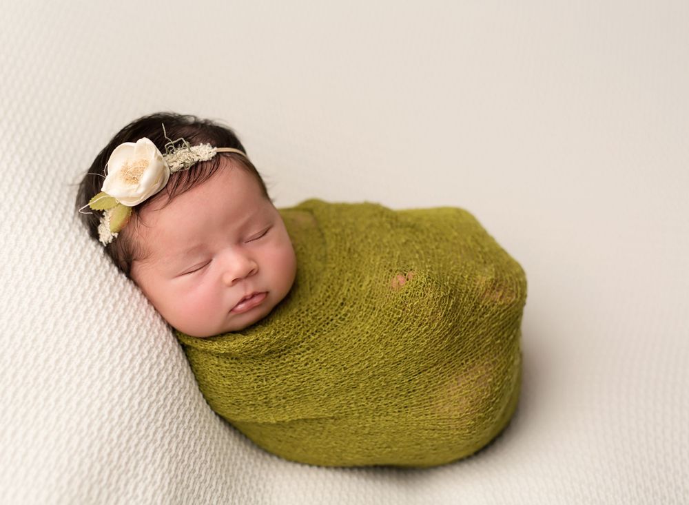 Sleeping newborn wrapped in olive green wrap with delicate white flower headband.