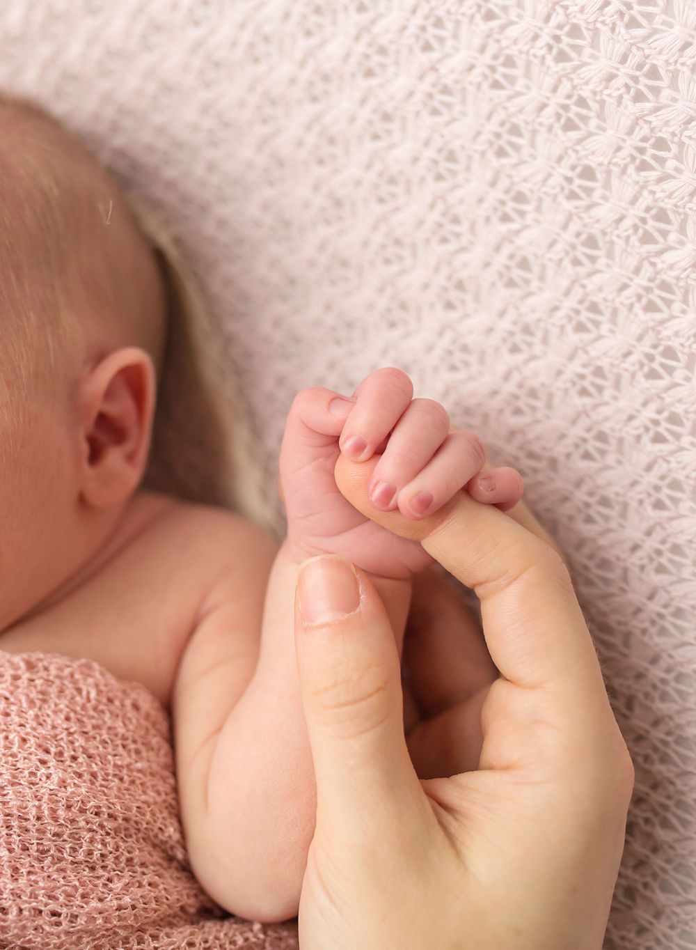 Tender moment of adult hand holding tiny newborn fingers against lace background.