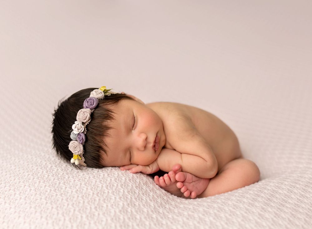 Peaceful sleeping newborn wearing delicate floral headband on white backdrop.