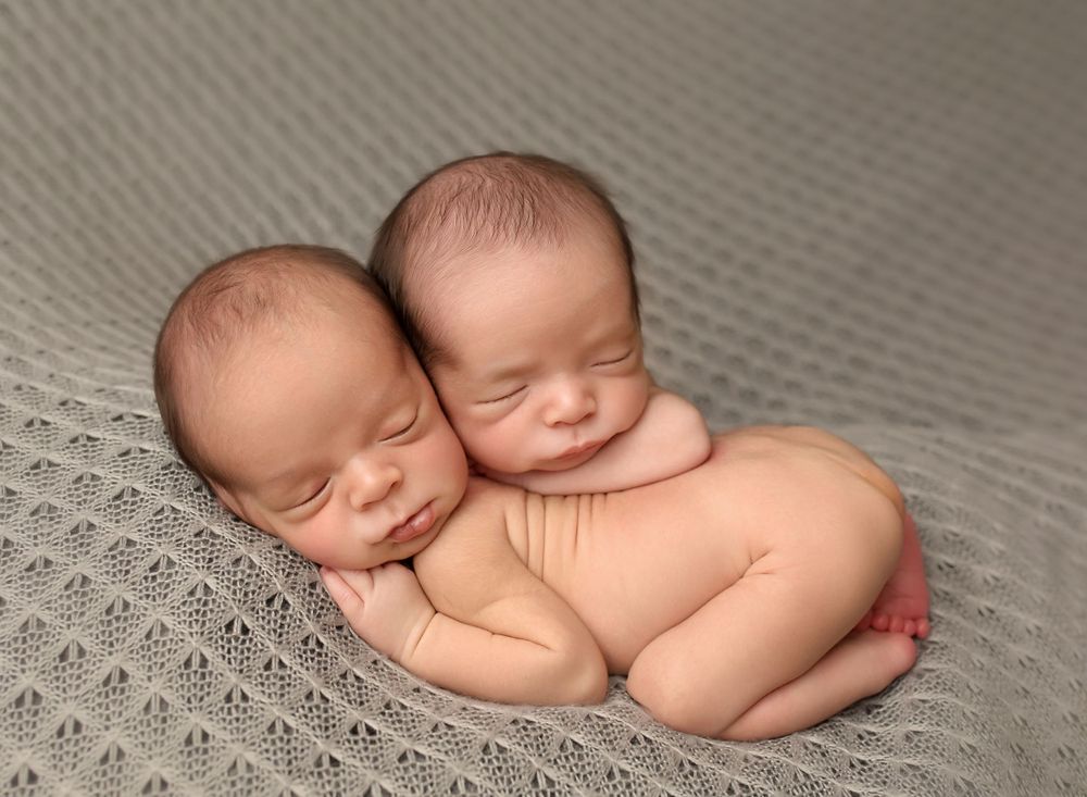 Peaceful sleeping newborn twins cuddled together on textured grey blanket.