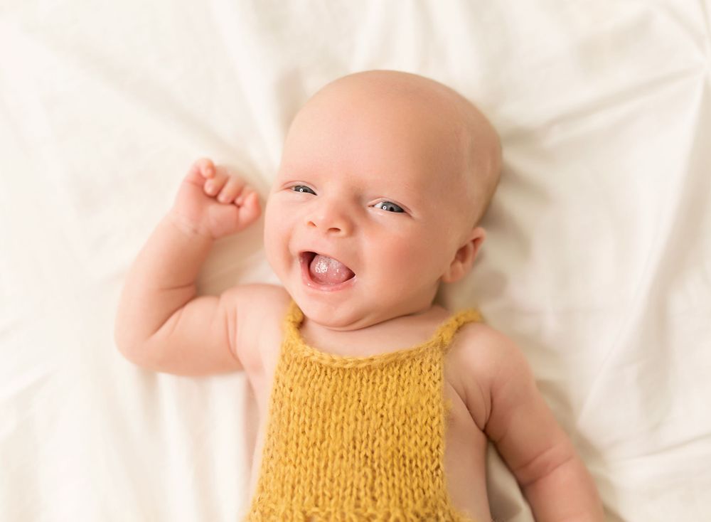 Smiling baby wearing yellow knit tank top lying on white bedding.