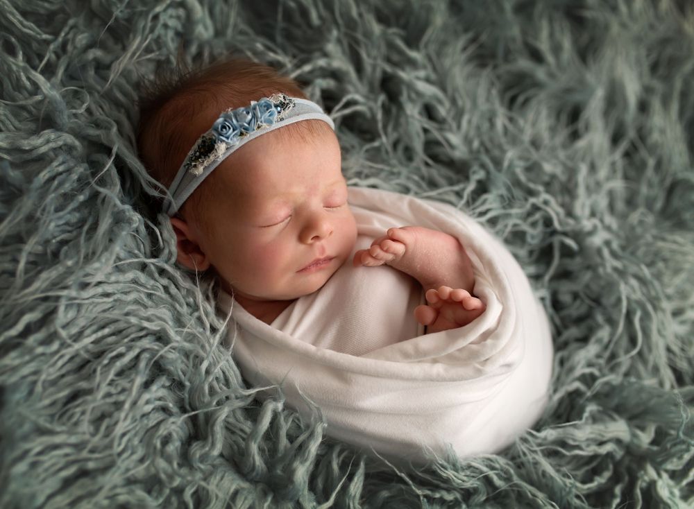 Sleeping newborn wrapped in white swaddle on grey fur with delicate headband.