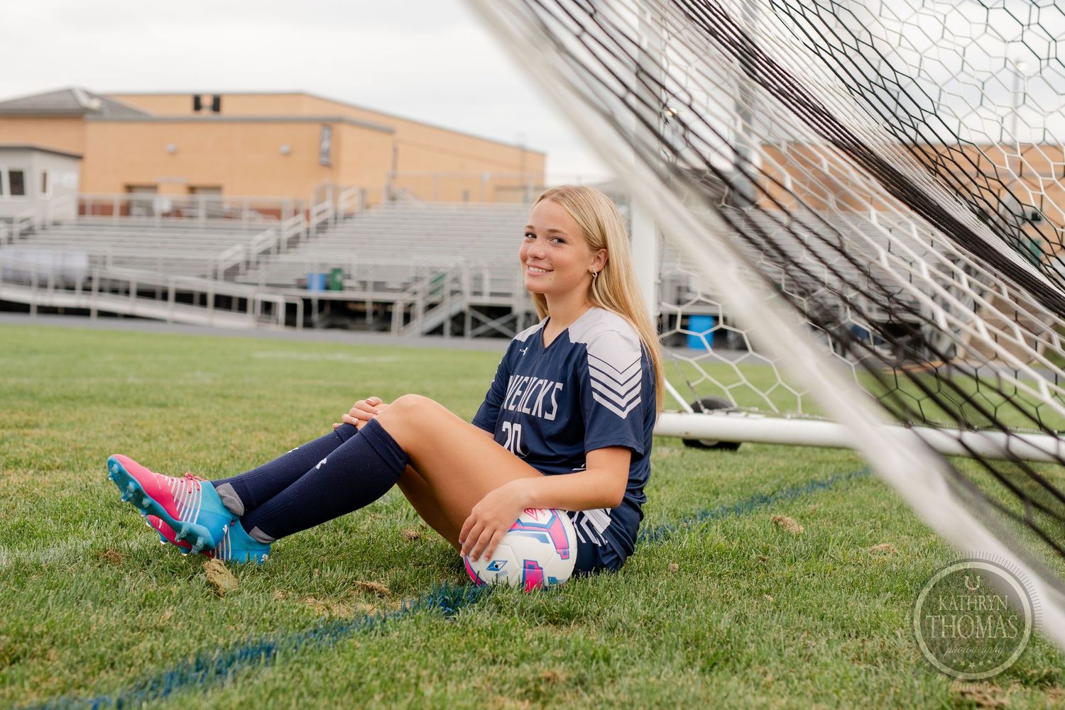Soccer player in navy jersey sitting by goal post with ball on field.