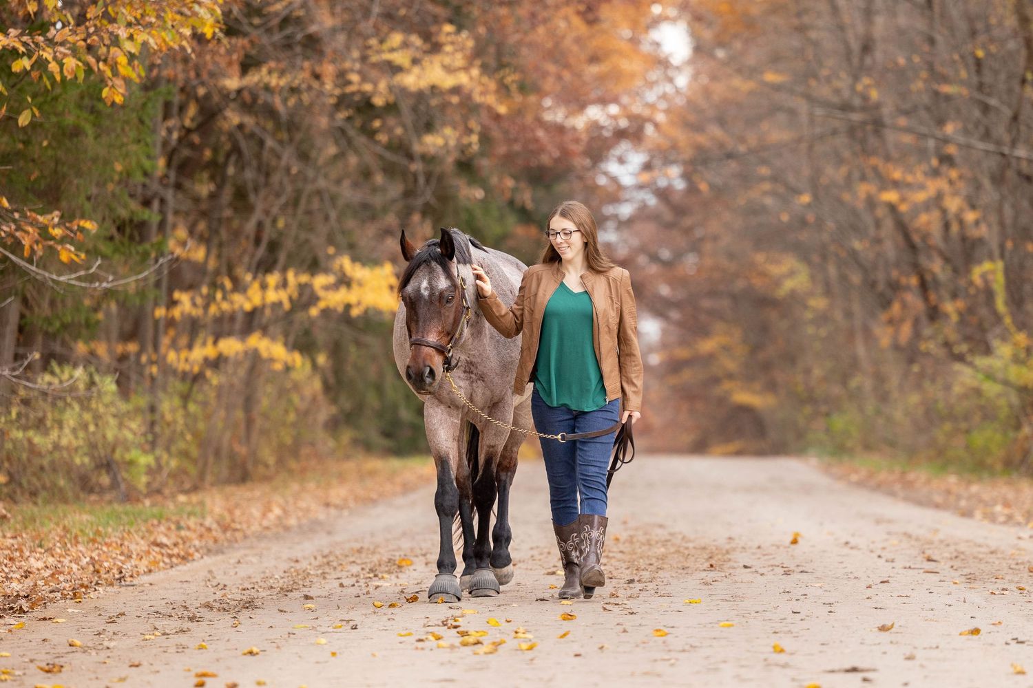 Senior photo of a girl in a green shirt and brown jacket in the fall with her bay roan horse walking down a road
