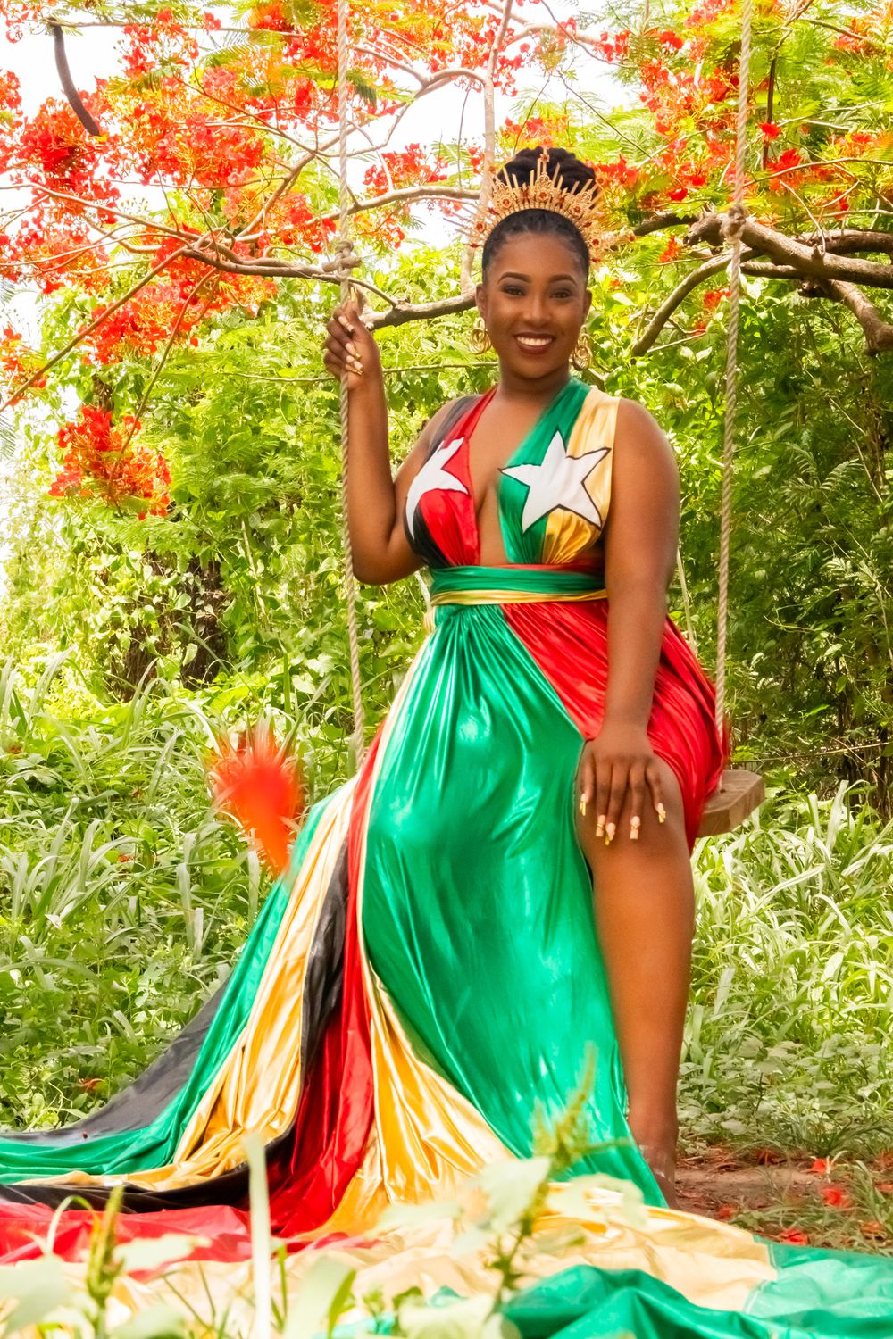 Smiling woman wearing crown and St. Kitts flag-inspired gown poses under red flowering tree outdoors.
