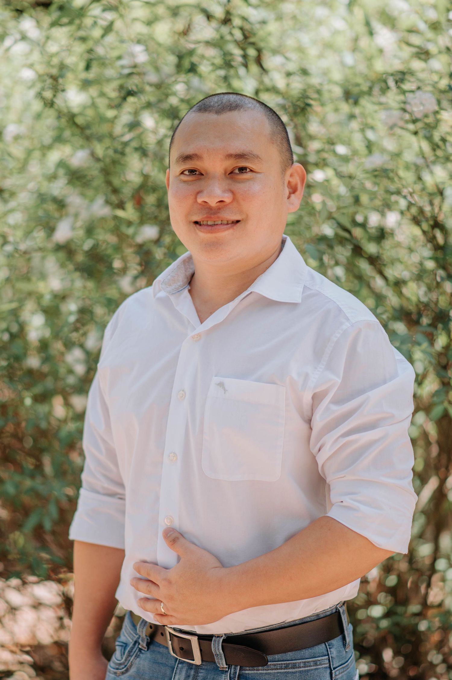 A man wearing a white button-down shirt and jeans poses for a casual portrait in a garden setting.
