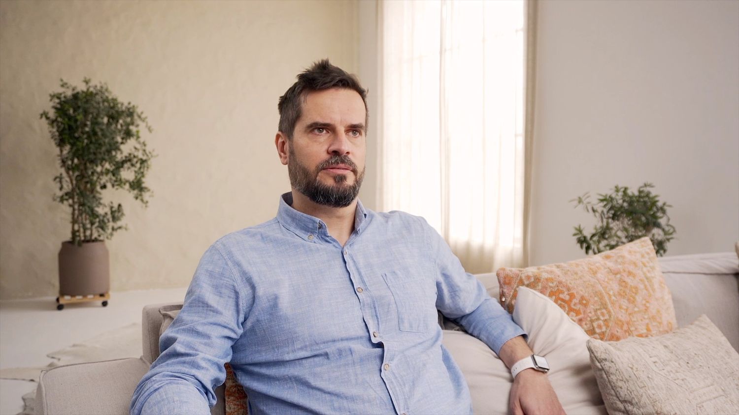 A person in blue shirt sits casually on a beige couch in a bright, modern living room.