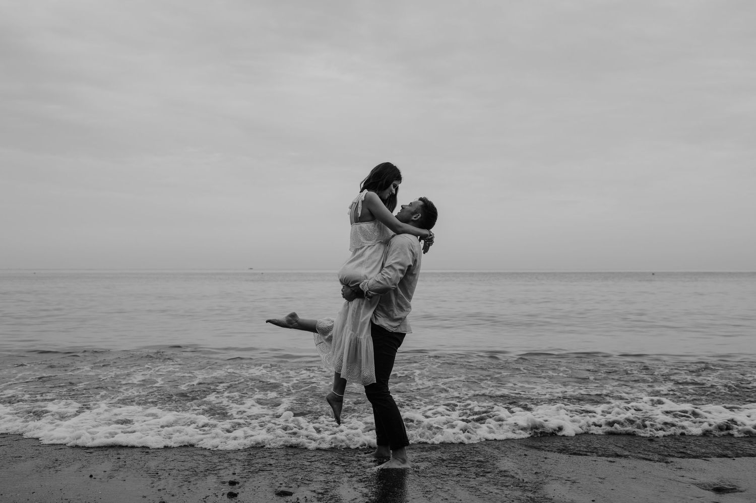 A couple embraces in a romantic moment by the ocean shore in black and white photography.