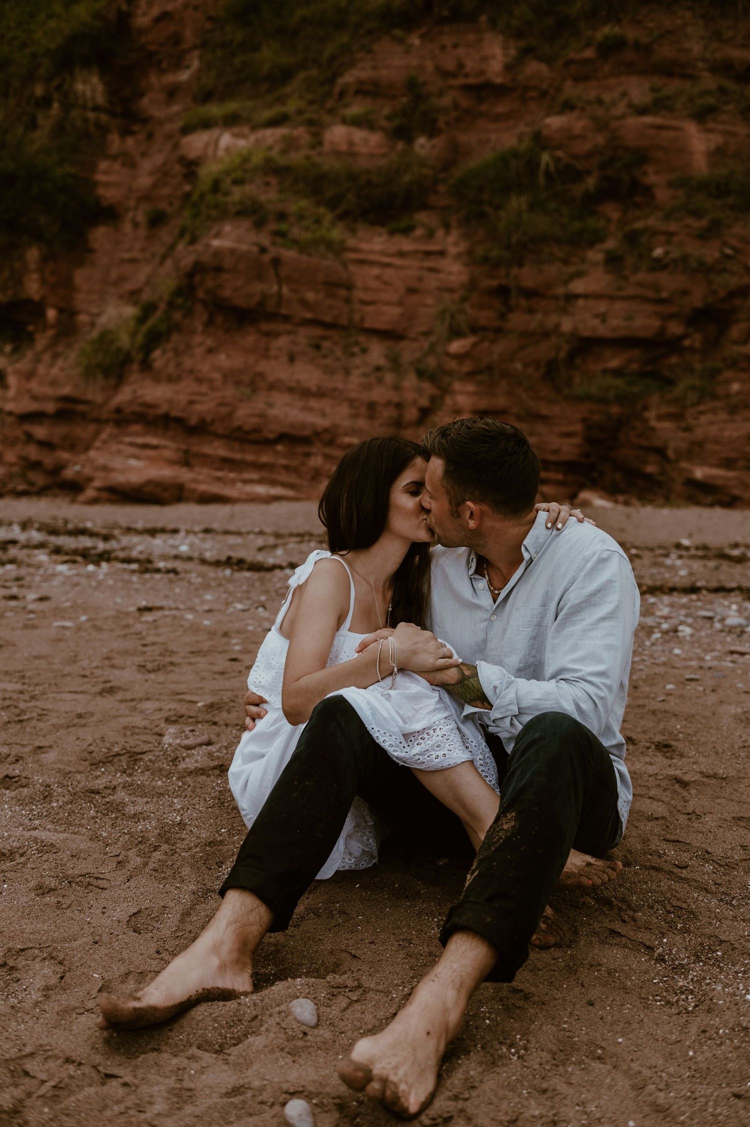 Romantic couple sharing an intimate moment on a beach with rustic red cliffs in the background at sunset.