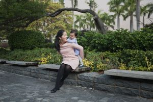 Parent and child sit together on stone wall surrounded by lush tropical garden with palm trees.