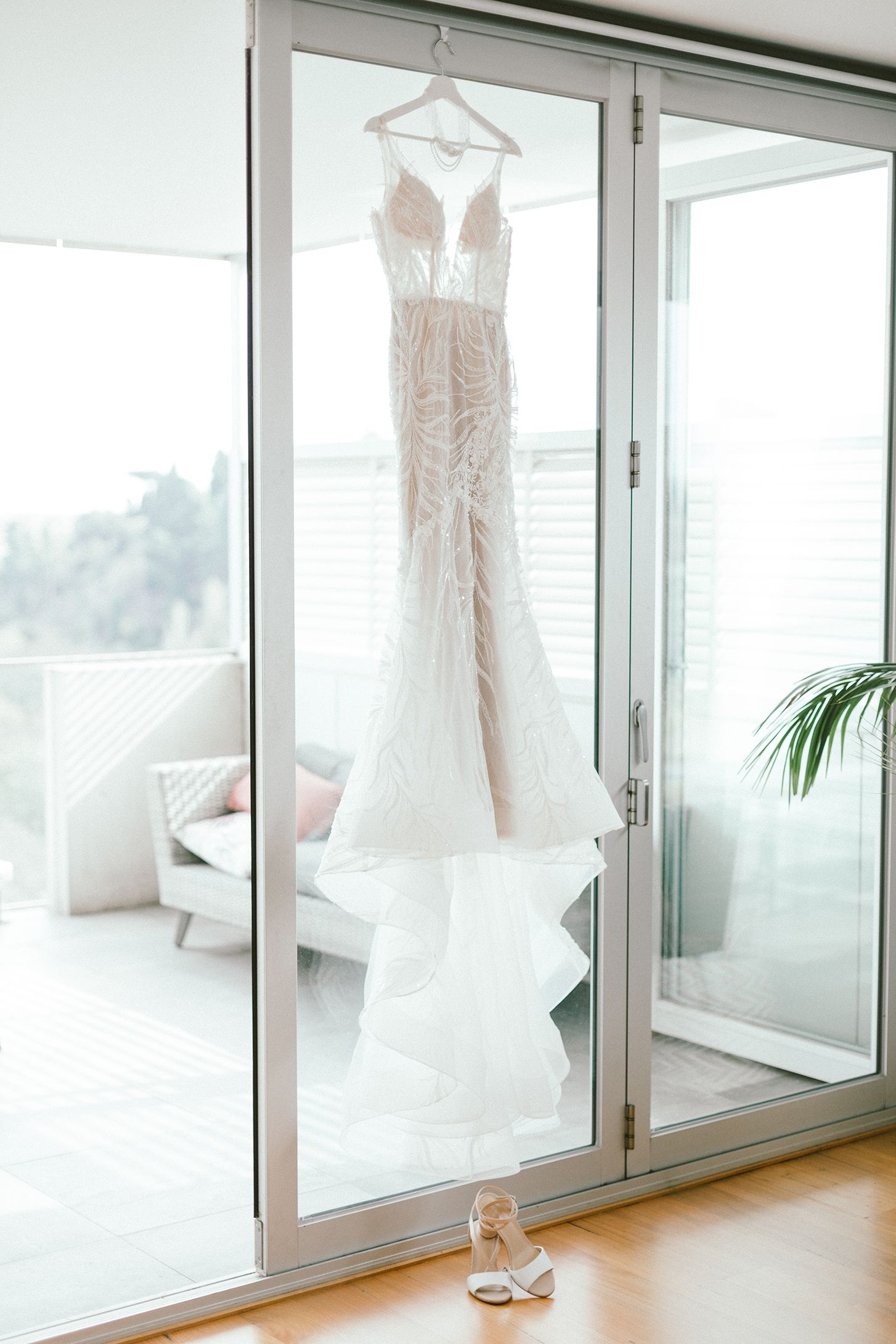 An elegant white wedding gown with lace detailing hangs against sliding glass doors with a pair of shoes below.