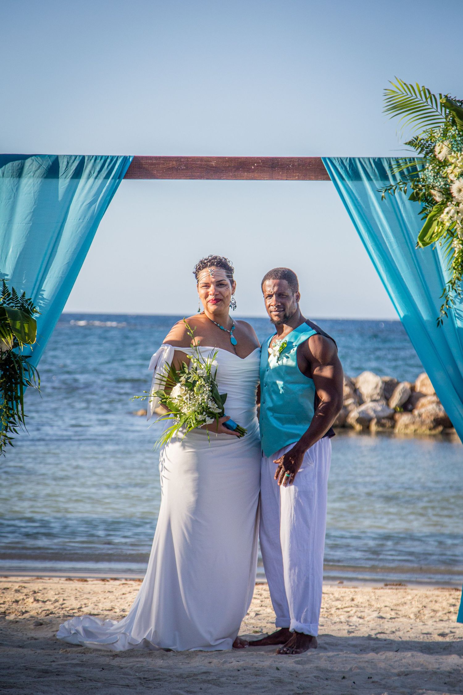 Newlywed couple posing under turquoise arch on beach with ocean backdrop.