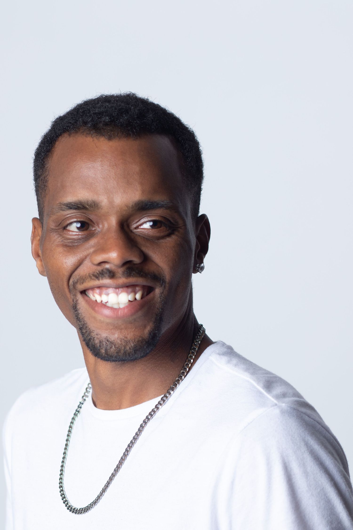 A studio portrait of a person in a white t-shirt and silver chain necklace against a light background.
