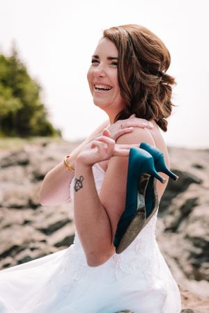 Joyful bride in white dress laughing on rocky beach.