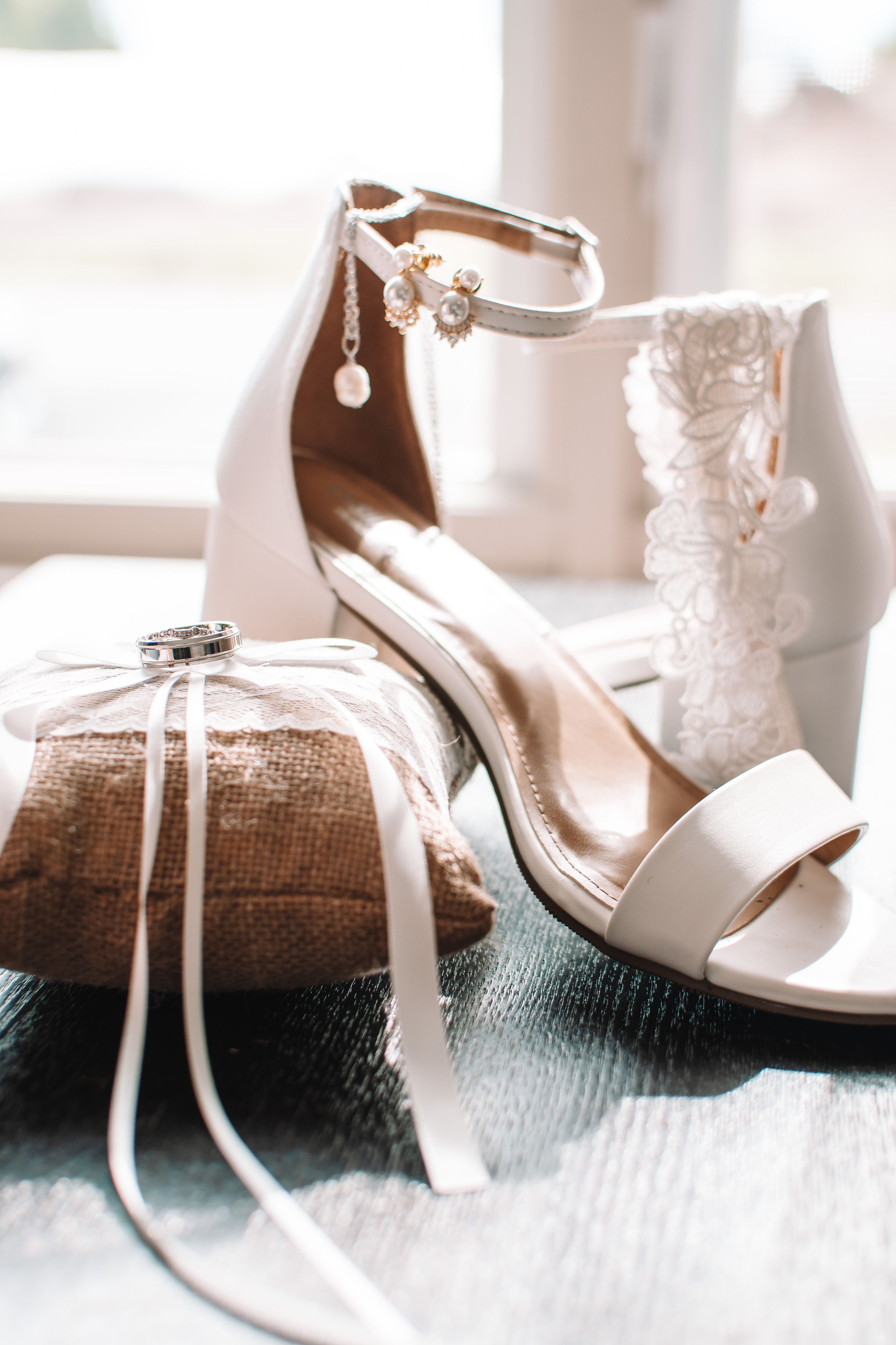 Elegant white bridal sandals with delicate ankle straps and wedding rings on textured surface.