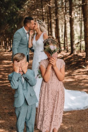 Playful wedding portrait in a forest with children covering their eyes in the foreground.