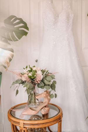Elegant white wedding dress hanging with bridal shoes and pink floral bouquet on rattan table.