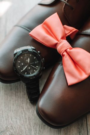 Black watch and coral bow tie arranged with brown leather shoes on wooden surface.