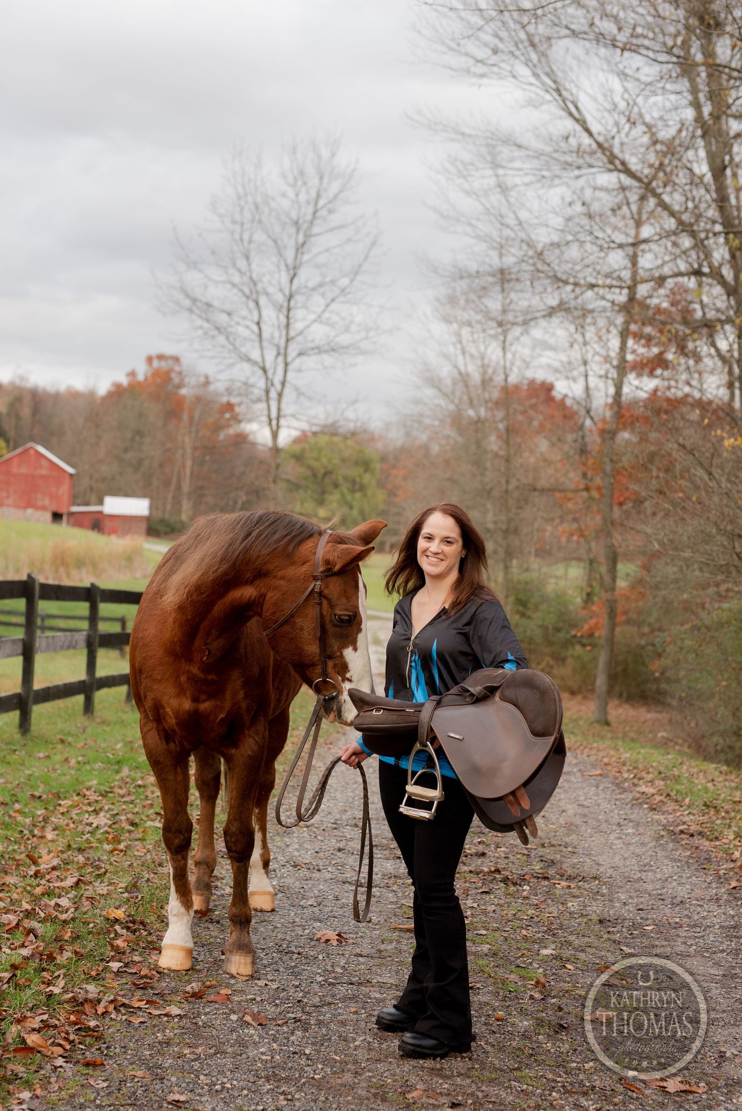 A chestnut horse stands with rider on a dirt path surrounded by autumn trees and black fencing.