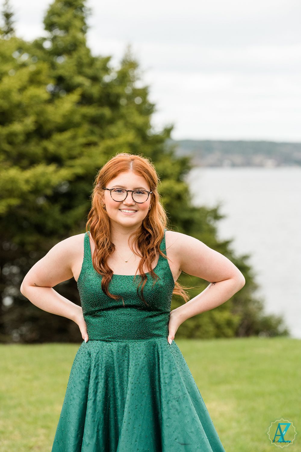Elegant emerald gown worn in outdoor portrait with water and evergreens in background.