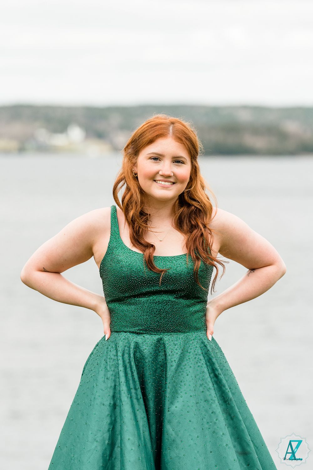 A person in an emerald green formal dress poses against a waterfront backdrop.