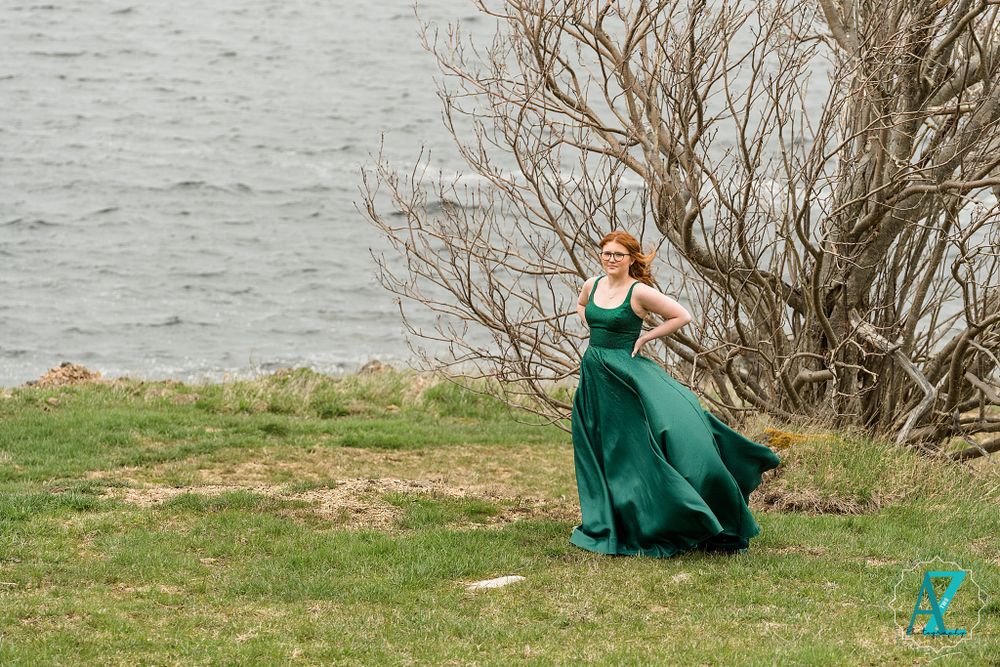 An elegant green dress flows in the wind by the waterfront near bare branches.