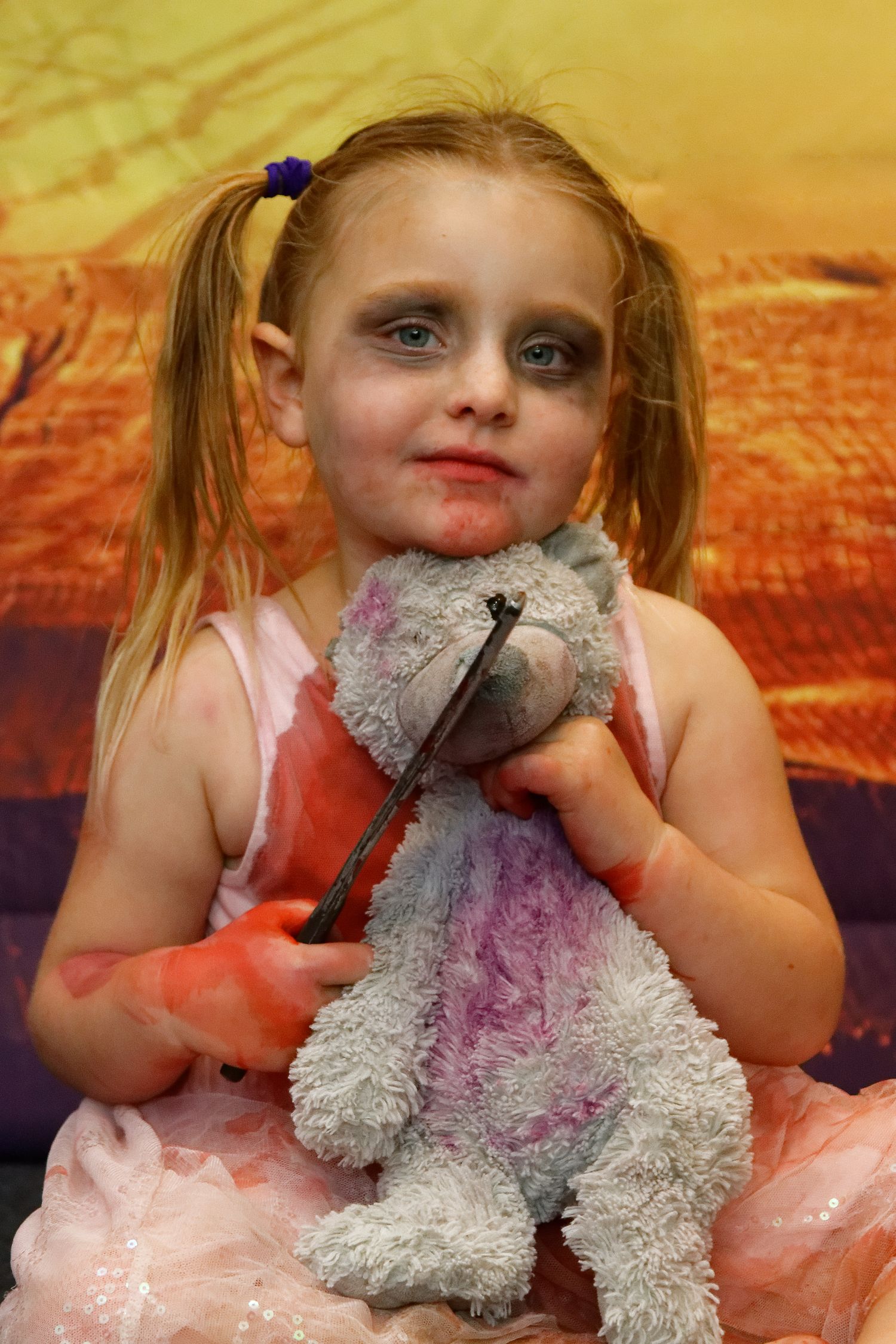 Halloween portrait with child in pink dress holding damaged teddy bear against orange backdrop.